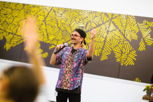 A man with a microphone in front of a graphic yellow painting, with raised arms in the foreground
