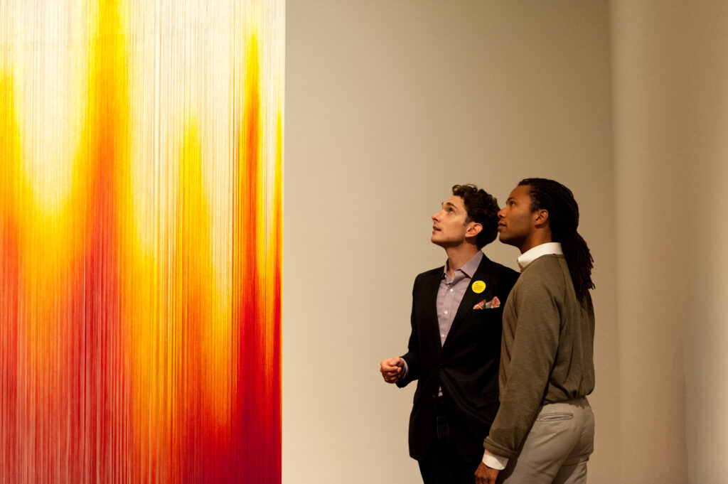 Two men look at a sculpture made of bright red and orange hanging string