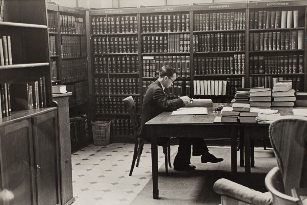 Black and white photo of a man in a suit seated at a table and looking through a card catalogue in a library