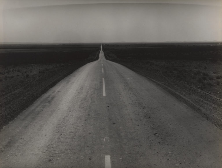 Black and white photograph of empty two-lane highway receding into the flat horizon with a cloudless sky above