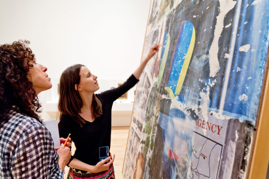 A woman points out part of a large painting to the woman next to her.