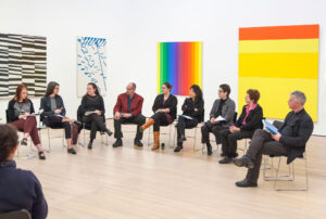 A group of people sitting in chairs arranged in a circle in a white gallery filled with colorful paintings