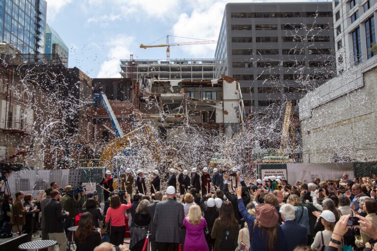 A crowd cheers and confetti flies as a line of people in suits and hard hats break ground at the SFMOMA construction site