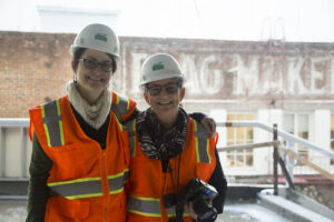 Two women in glasses, hard hats, and bright orange constrction vests stand arm in arm