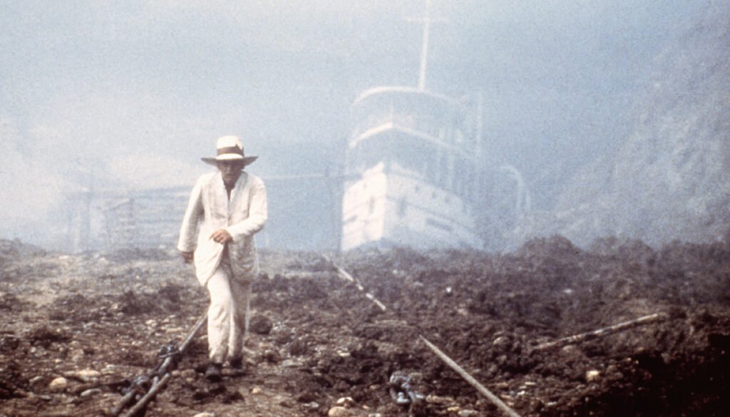 A film still of a man in a white suit and hat walking across a desolate landscape