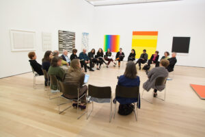 A group of people sitting in chairs arranged in a circle in a white gallery filled with colorful paintings