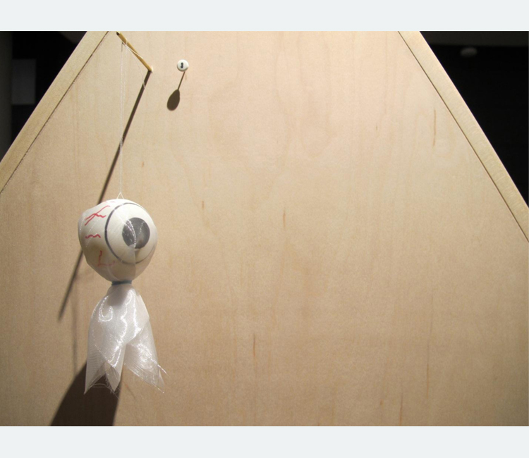 Plastic eyeball wrapped in translucent white fabric hanging off a dowel on a wooden board