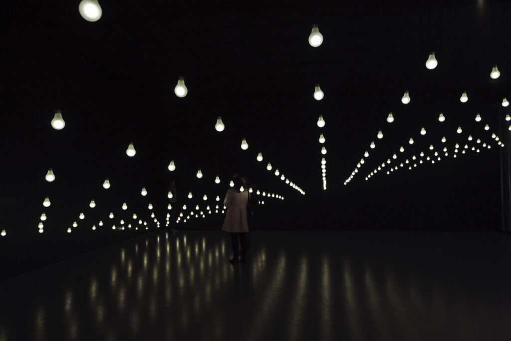 A visitor stands in a darkened room with white light bulbs running the length of the 