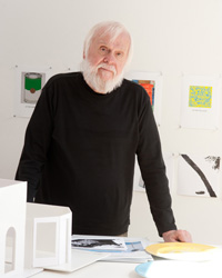 A man with white hair and a beard stands behind a table in front of a wall hung with works on paper