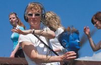 A woman stands against a blue sky wearing headphones connected to microphones on the ears of the blue mannequin head she is holding