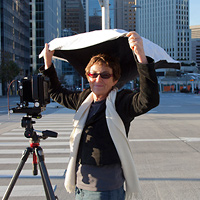 A woman stands behind a camera mounted on a tripod and holds a piece of fabric over her head to block the light