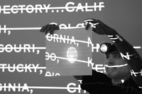 A black and white photograph of a man wearing a headlamp holding up a piece of paper on which text and lines are being projected