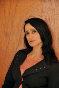 A woman in a black jacket leans against a wooden wall