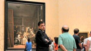In a gallery, a man with folded arms looks into the camera as he stands in front of a Renaissance painting.