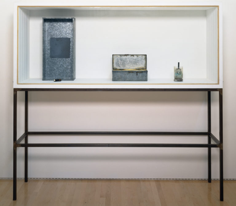 A white-walled vitrine atop thin steel legs, containing several metal and glass objects