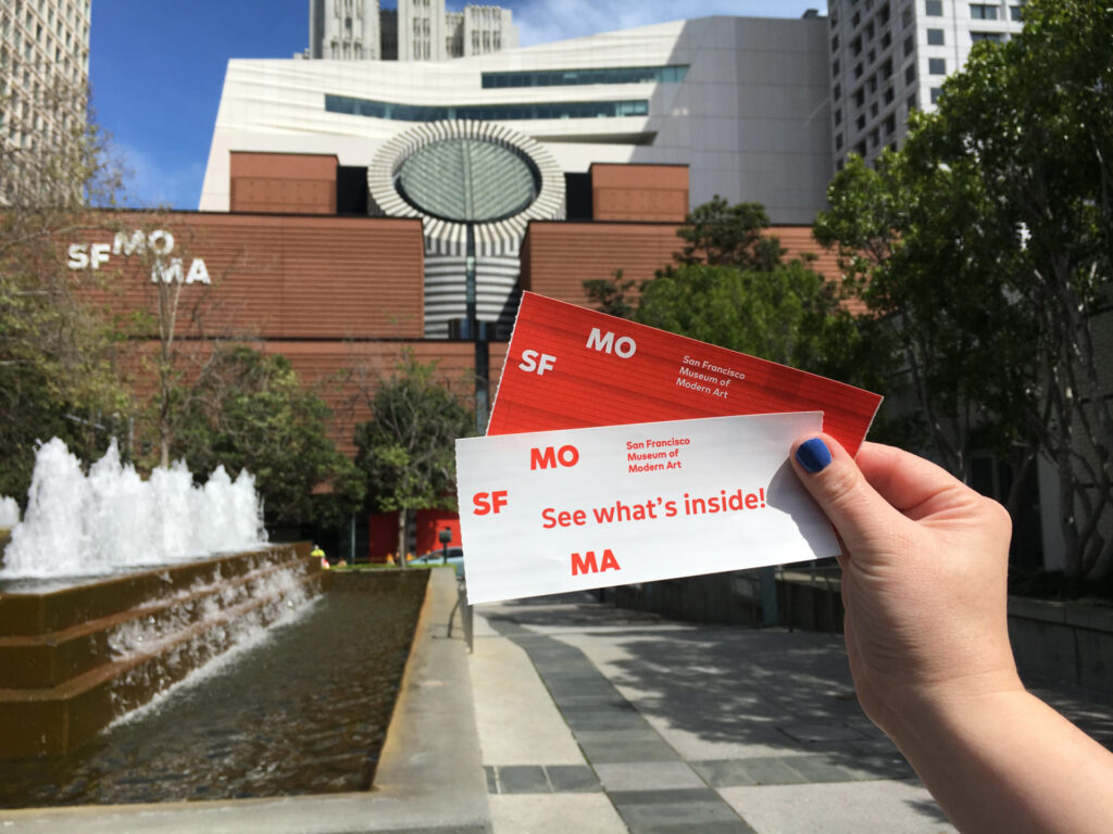 A disembodied hand holds two tickets in front of the new SFMOMA building