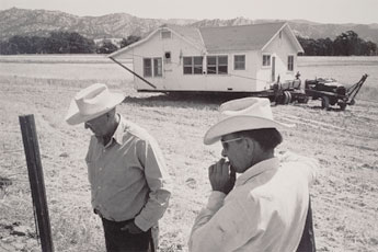 two men in cowbow hats on farm with house in back