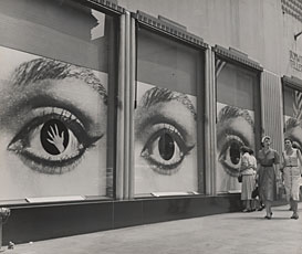 three large city windows covered with image of woman's eye; two women walking by windows in corner