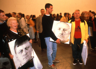 Gerz, crowded room with two people holding images of men's portraits