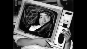 A black-and-white photograph of artist Nam June Paik with his face poking through a screen-less TV 