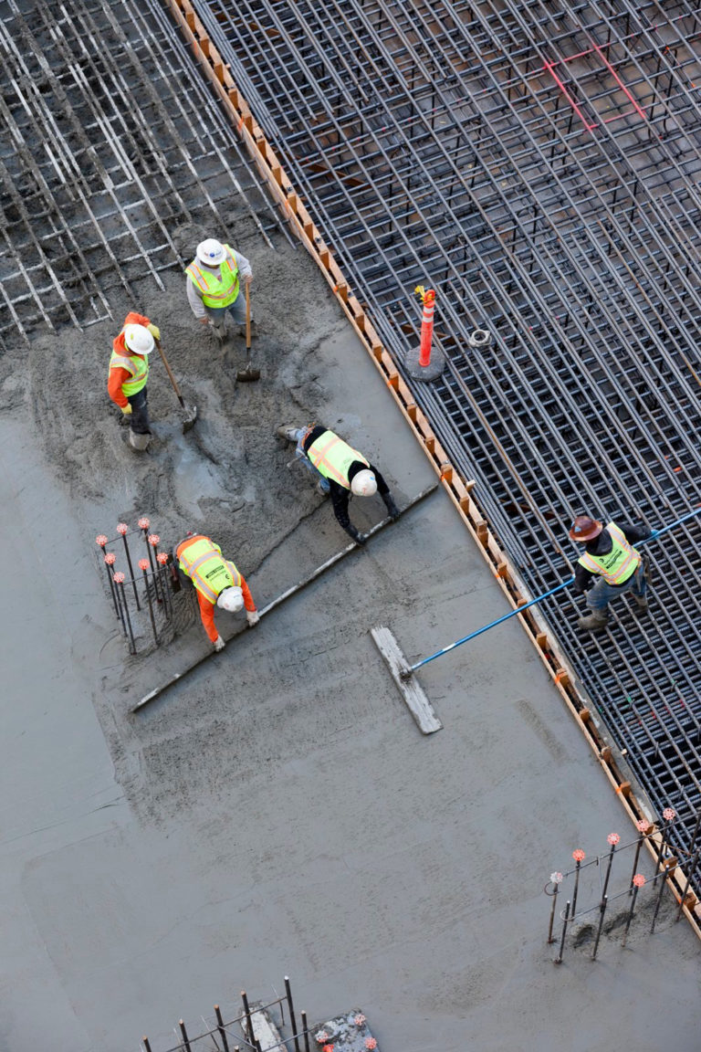 An overhead shot shows five construction workers laying a concrete foundation