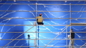 Artists standing on scaffolding work on a large wall painting of white wavy lines on a bright blue blackground