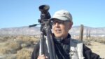 Photographer Anthony Hernandez holds a camera on a tripod over his shoulder, with a view of the Mojave Desert behind him.