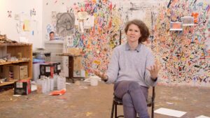 Artist Dana Schutz in her studio, sitting in front of a colorful-paint-splattered wall