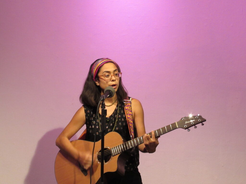 A woman with a guitar stands in front of a pink wall