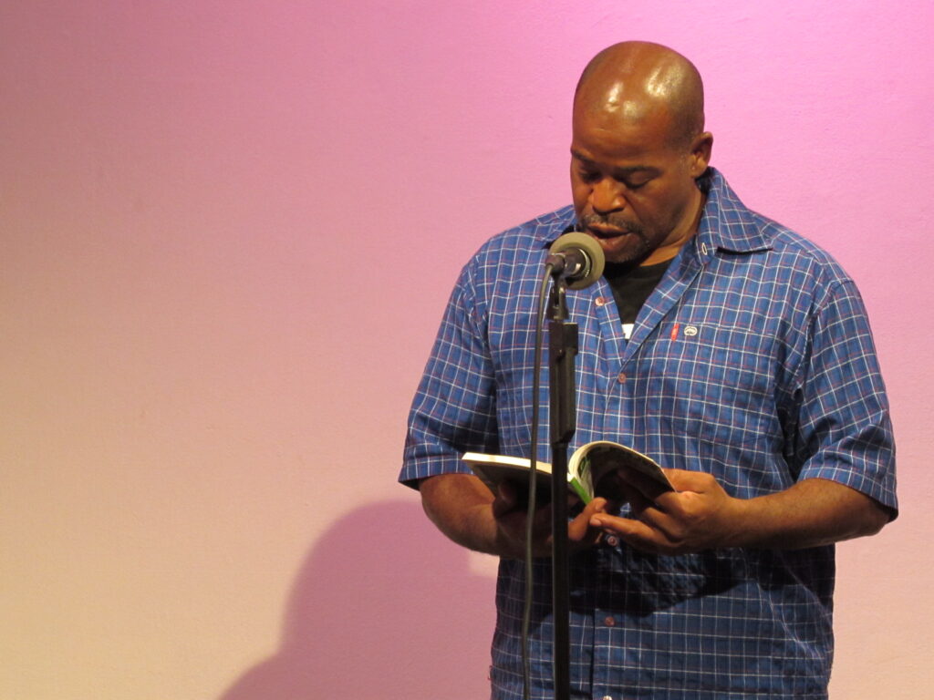 A man stands in front of a pink wall reading from a book
