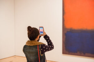 A woman in a bun holds her cell phone up to take a picture of an orange and blue painting