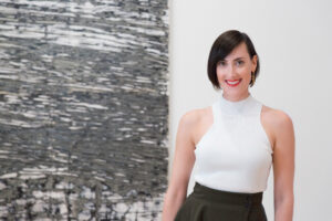 A woman wearing a white top and black skirt stands in front of an artwork