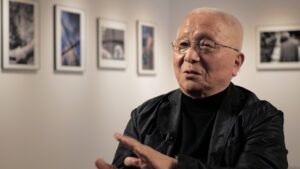 Photographer Kikuji Kawada in a photo gallery, with photographs out of focus in the background