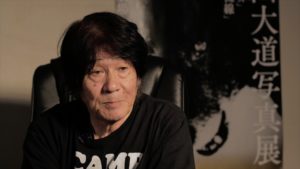 Photographer Daido Moriyama wearing a shirt reading "CAMP," sitting in front of a print of a wolf.