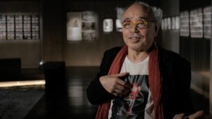 Photographer Nobuyoshi Araki in a photo gallery, wearing a shirt with a monster on it