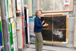A woman stands before a black encaustic painting hanging on a storage wall