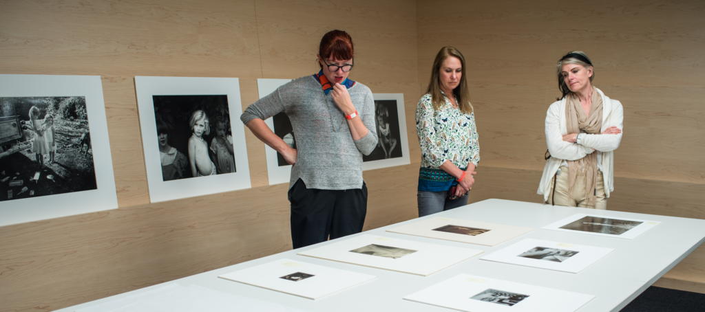Three Caucasian women stand over a large white table looking at black and white phtoographs