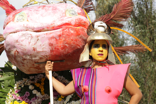 A woman with yellow facepaint, a gold helmet and a futuristic suit made of indigenous woven textiles holds a giant papier-machie axolotl head outdoors