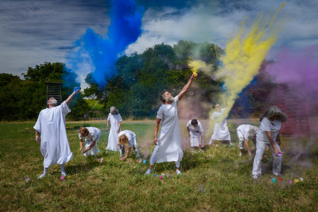 A group of adults wearing white togas fling colored powder into the air while standing in a field on a sunny day