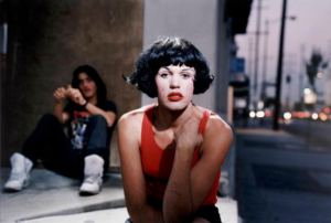 A heavily made-up figure wearing a black wig and red lipstick sits on an urban stoop with a young man visible behind