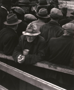 Black and white photograph, shot from above, of downtrodden men in hats and dark coats standing in line