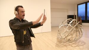 A man holds a smartphone up ahead of him in a gallery, with a metal sculpture behind him