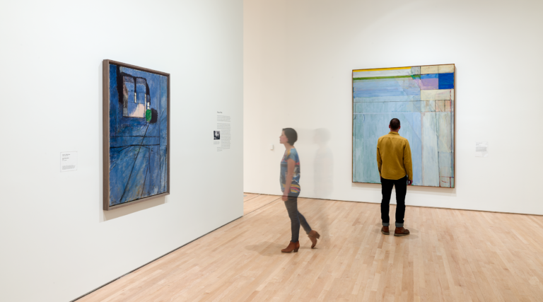 A woman walks toward a blue painting while a man stands in front of a large tranquil painting