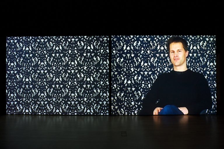 An image of a man sitting in front of a patterned wall.