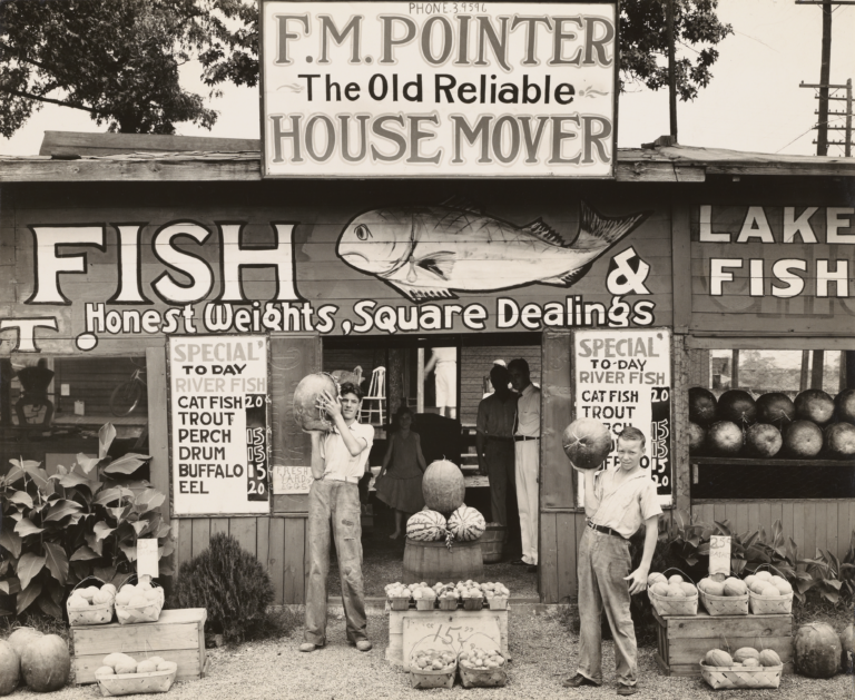 Two boys holding watermelons stand outside of a stand with a rooftop sign reading "F.M. Pointer - The Old Reliabe - House Mover" and the front wall painted with a fish and the words "FISH: Honest Weights, Square Dealings"