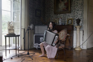 A woman in headphones sits alone playing an accordion in an ornately decorated but slightly ramshackle drawing room