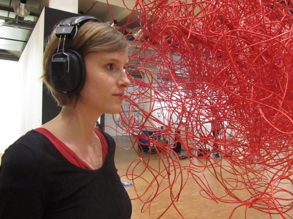 A woman in headphones stands listening next to a tangled, hanging mass of red cables