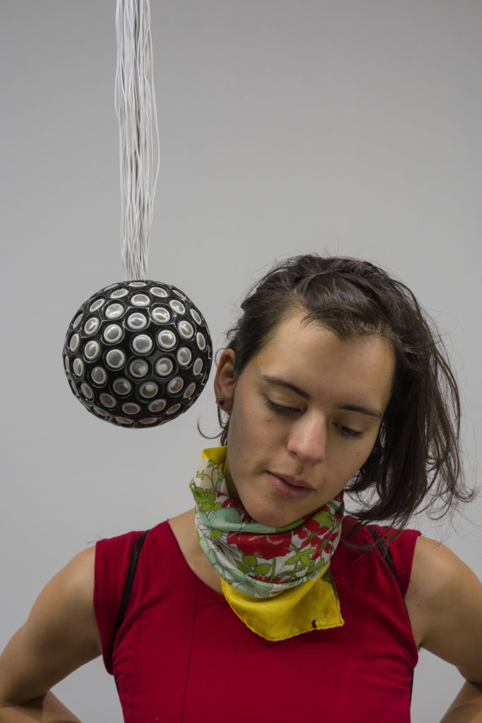 A woman stands looking down with her ear right against a hanging black orb covered in small silver speakers
