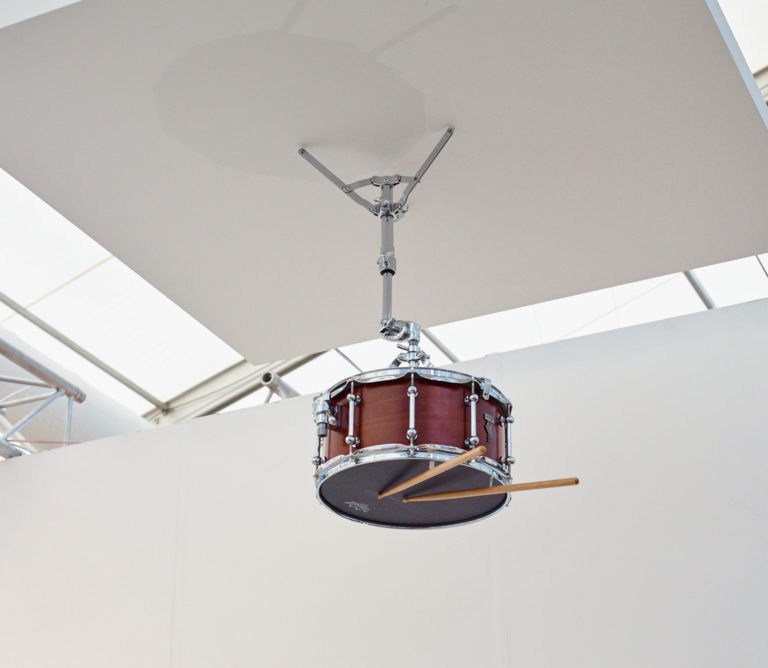 A single snare drum and two drumsticks are suspended from a ceiling panel 