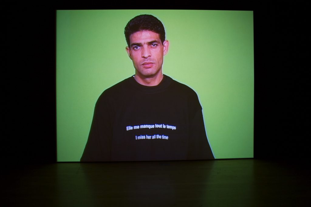 An image of a man in front of a green wall wearing a sweatshirt that says "I miss her all the time" in both English and French